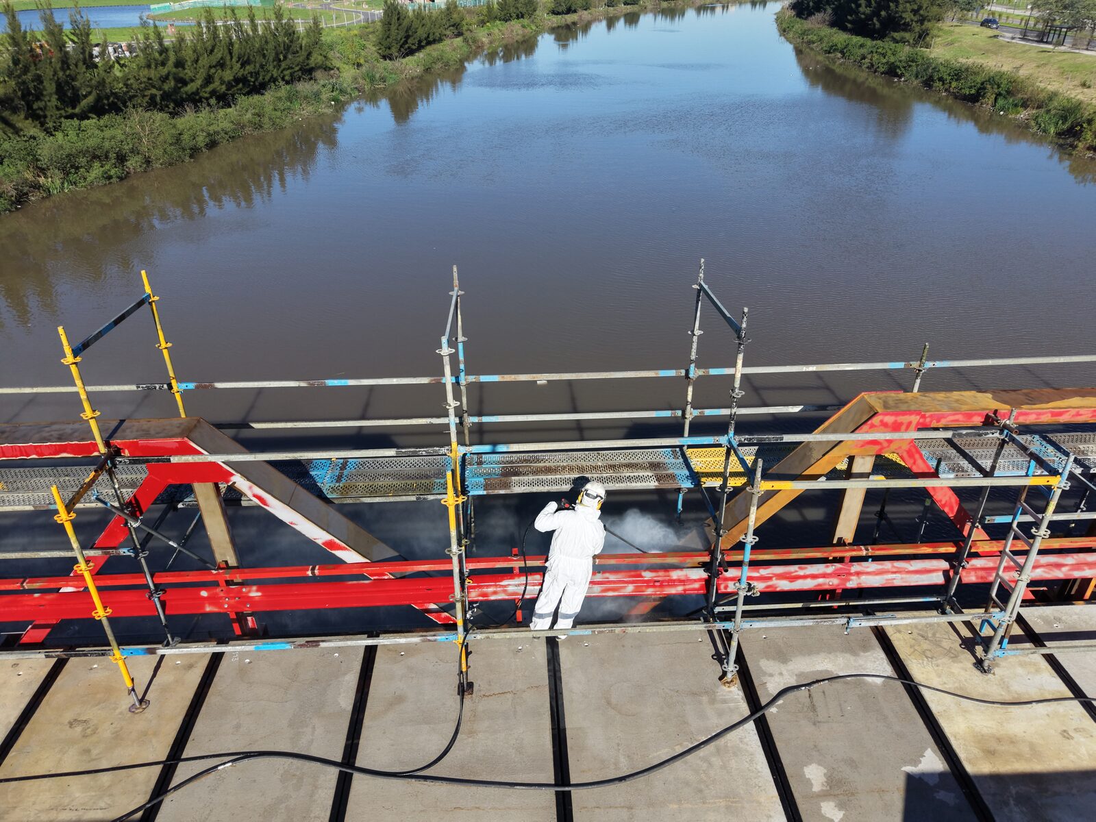 Operario con equipo de protección completo arenando el entramado metálico del puente desde un andamio, sobre el arroyo.